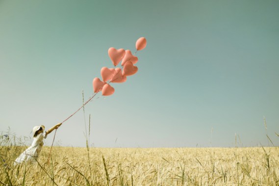 Young romantic girl with red heart balloons walking in a field of wheat.